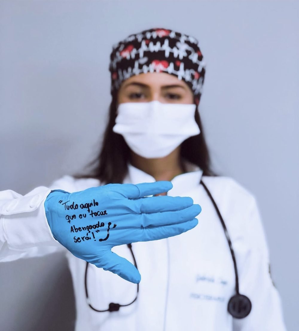 Gabriela Chagas de jaleco e estetoscópio, com a mão estendida em luva azul